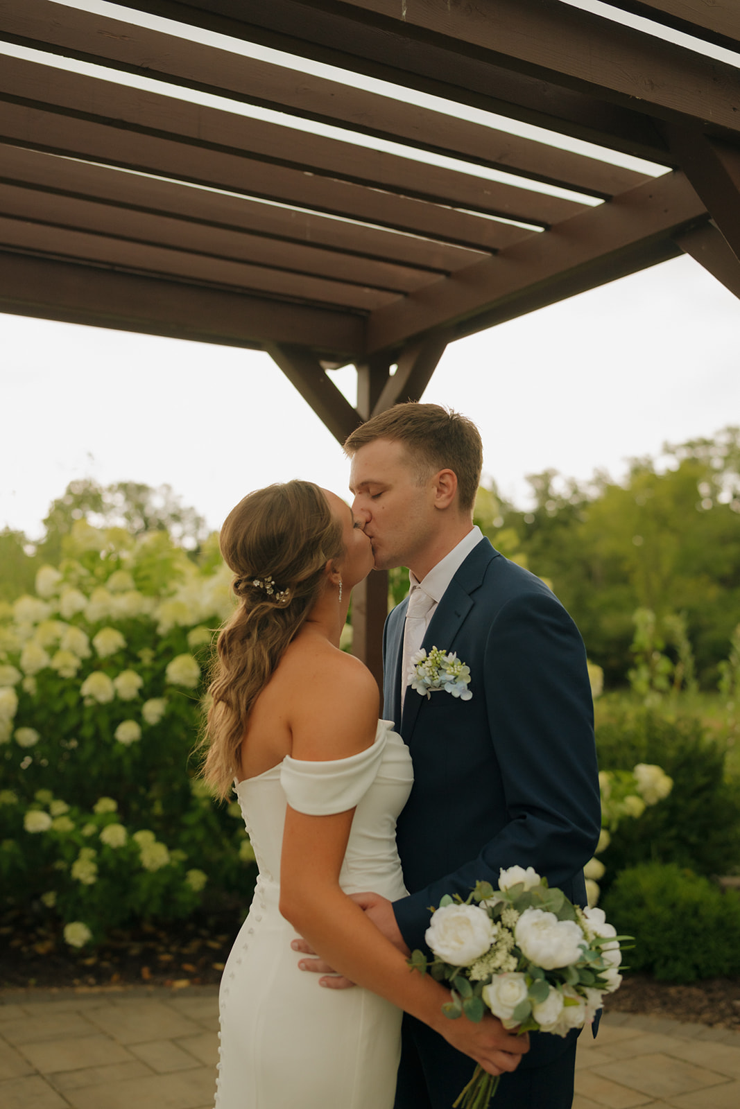 Close-up of a kiss under a wooden pergola, with the bride holding a soft white bouquet.