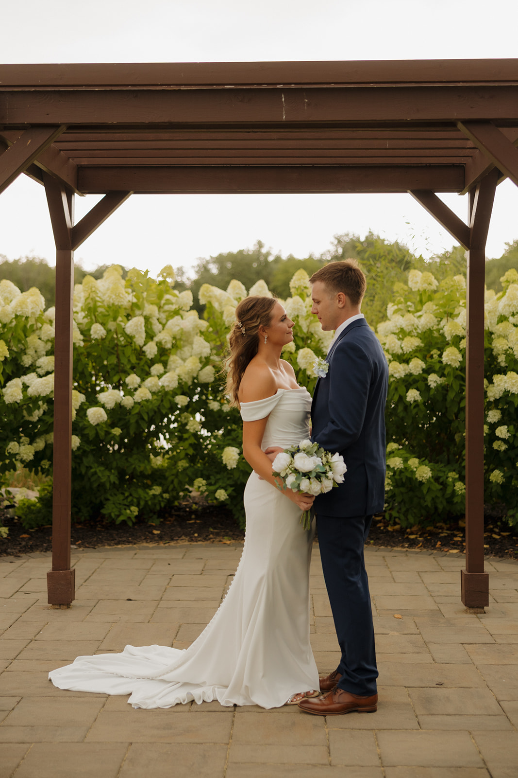 Couple standing beneath a wooden pergola surrounded by white hydrangeas, holding hands and smiling.