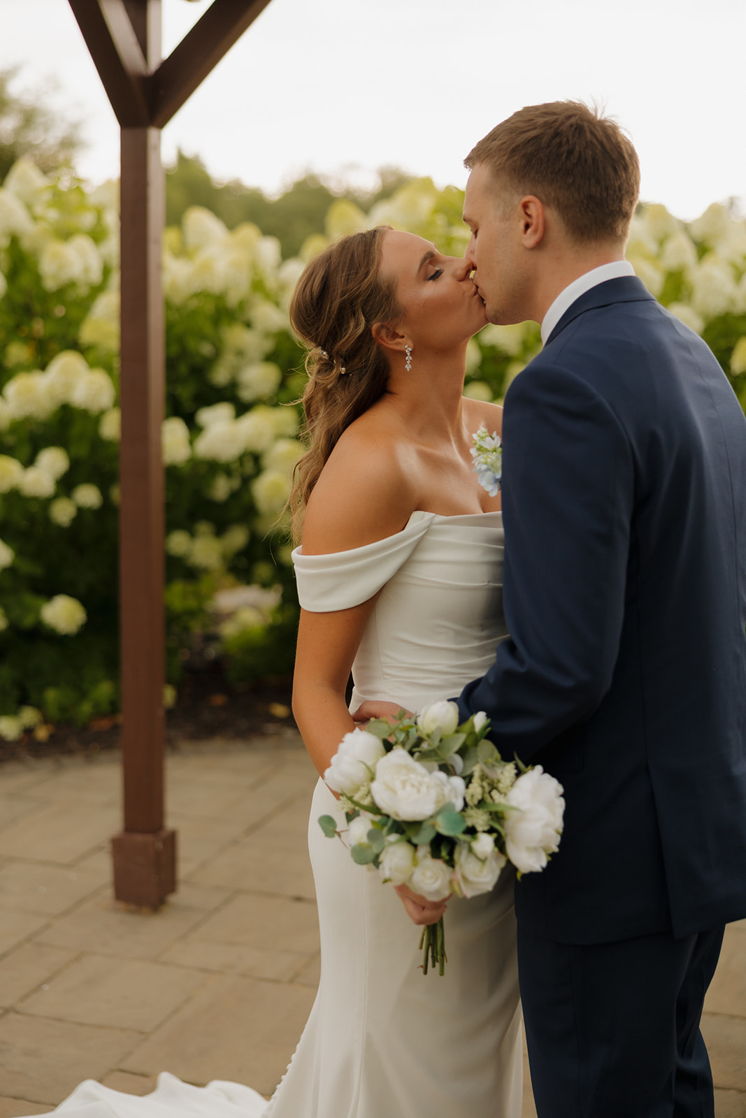 Bride and groom kiss under a wooden pergola, surrounded by blooming white hydrangeas.