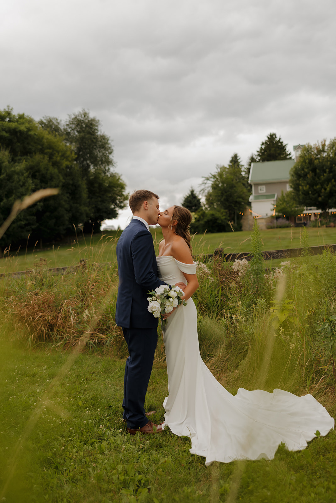 Bride and groom share a kiss in a grassy field with a barn in the distance at a romantic Wisconsin wedding venue.