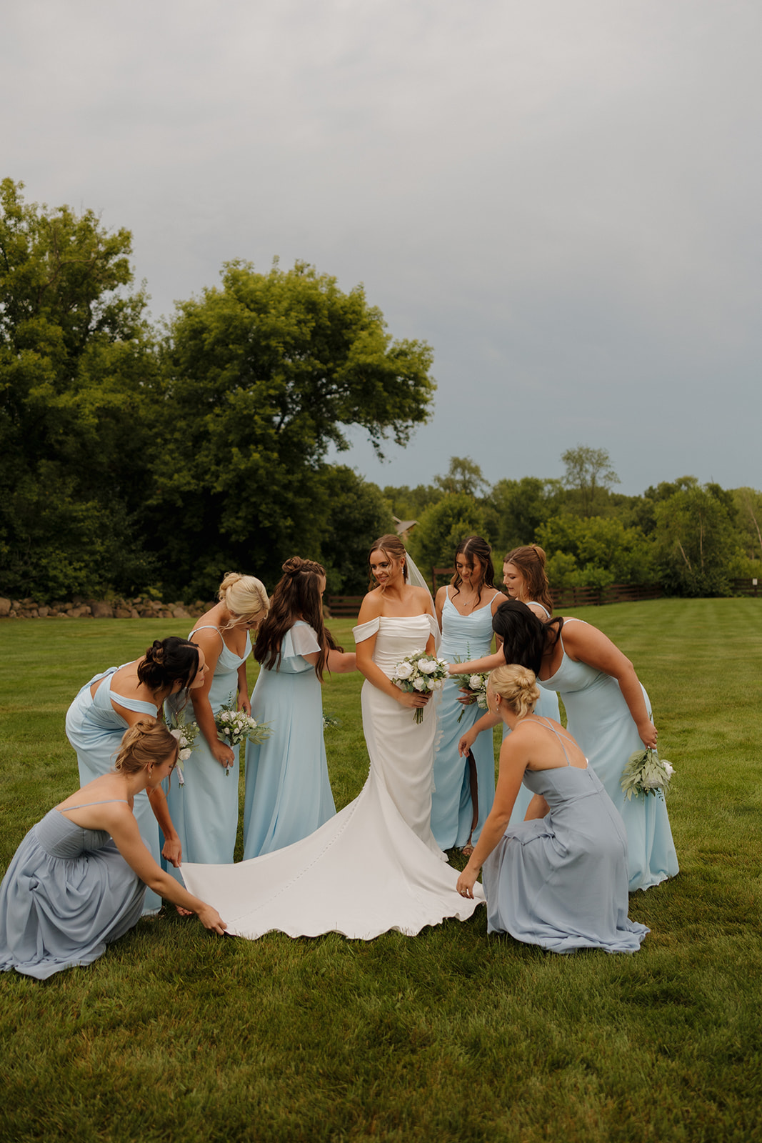 Bride standing on a lawn with her bridesmaids as they fan out the train of her dress.