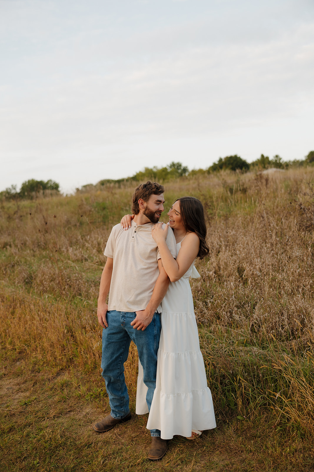 Engaged couple smiling at each other while wrapped in an embrace in a golden-hour field near Madison WI.