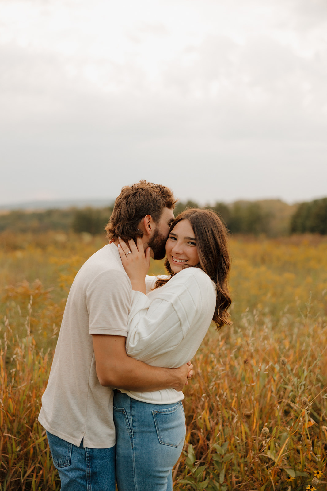 Smiling woman hugs her partner in a golden Madison WI field as he whispers in her ear.