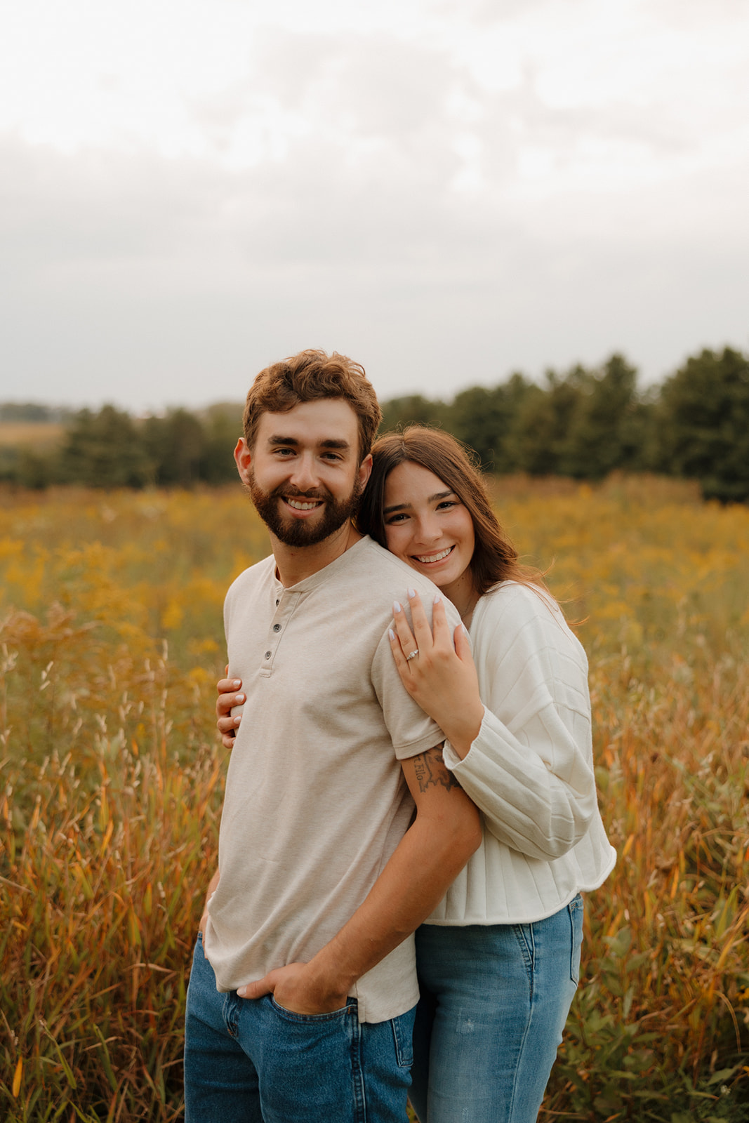 Engaged couple smiling in a sunlit Madison WI field, her hand resting on his shoulder to show off her ring.