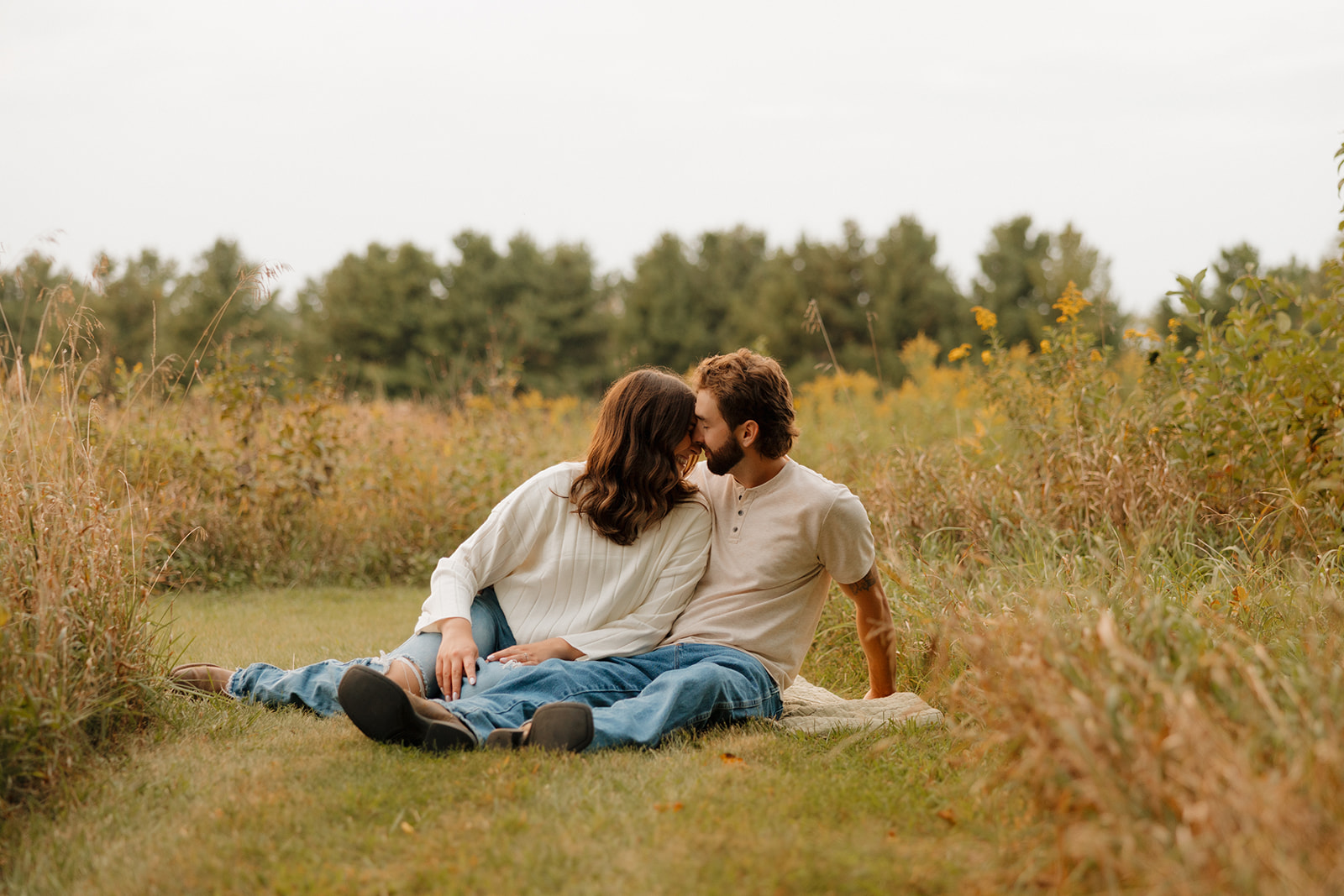 Engaged couple sitting on a blanket in a wide-open Madison WI field, sharing a quiet kiss.