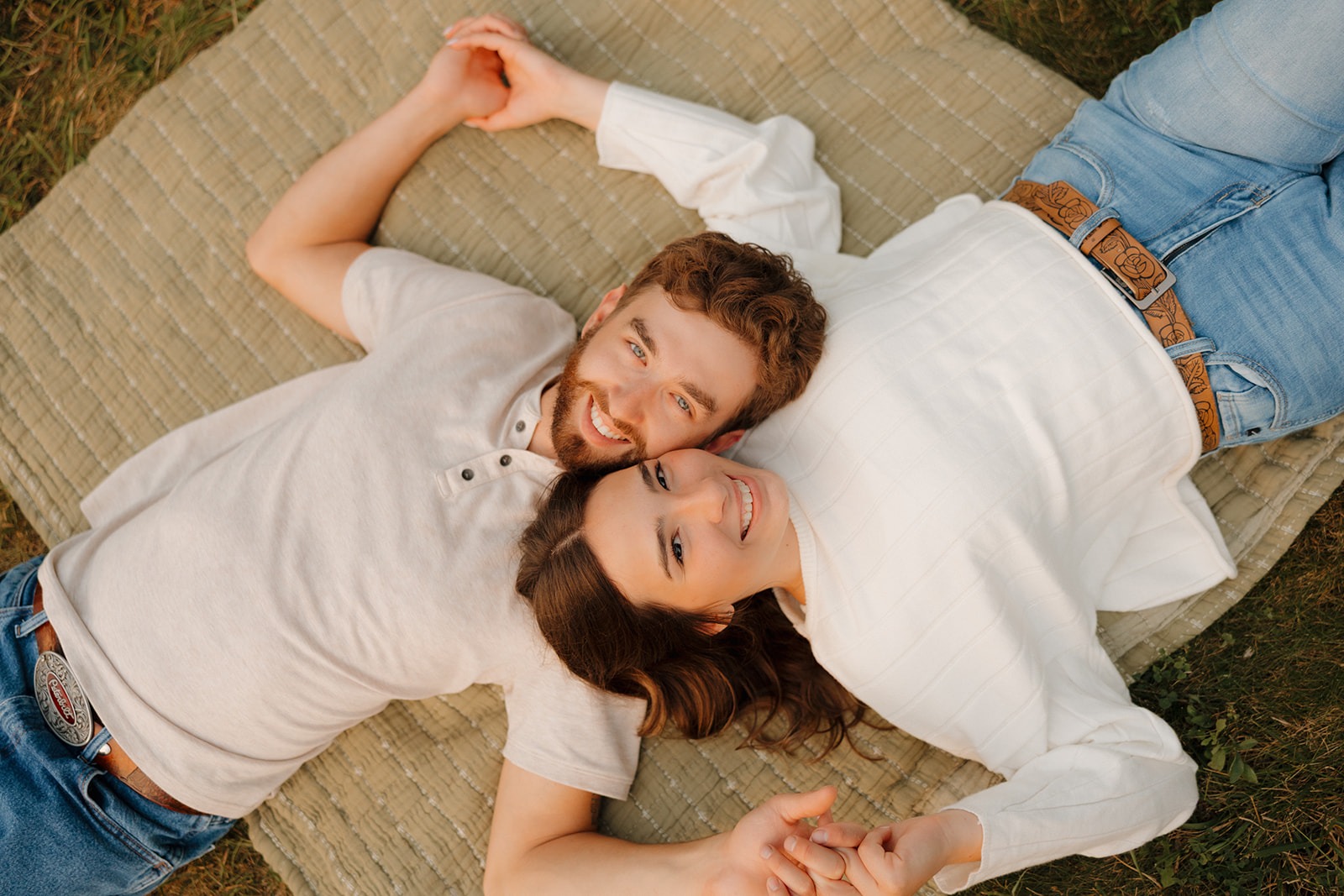 Overhead view of a couple lying side-by-side on a blanket in a Madison WI field, smiling up at the camera.