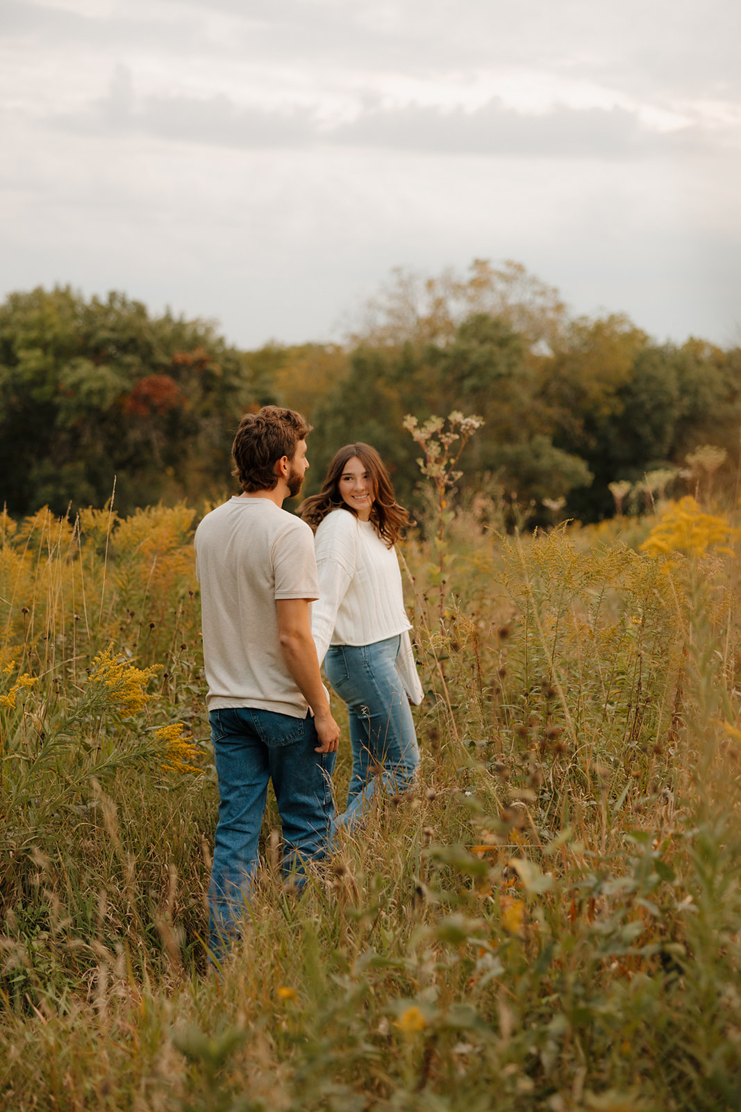 Couple walking through a lush field near Madison WI, turning back to exchange a glance and a soft smile.