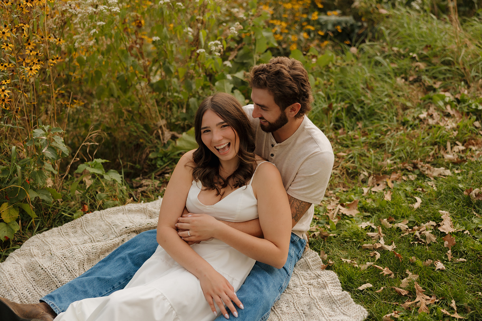 Couple laughing on a cozy blanket surrounded by wildflowers in a Madison WI meadow during their engagement session.