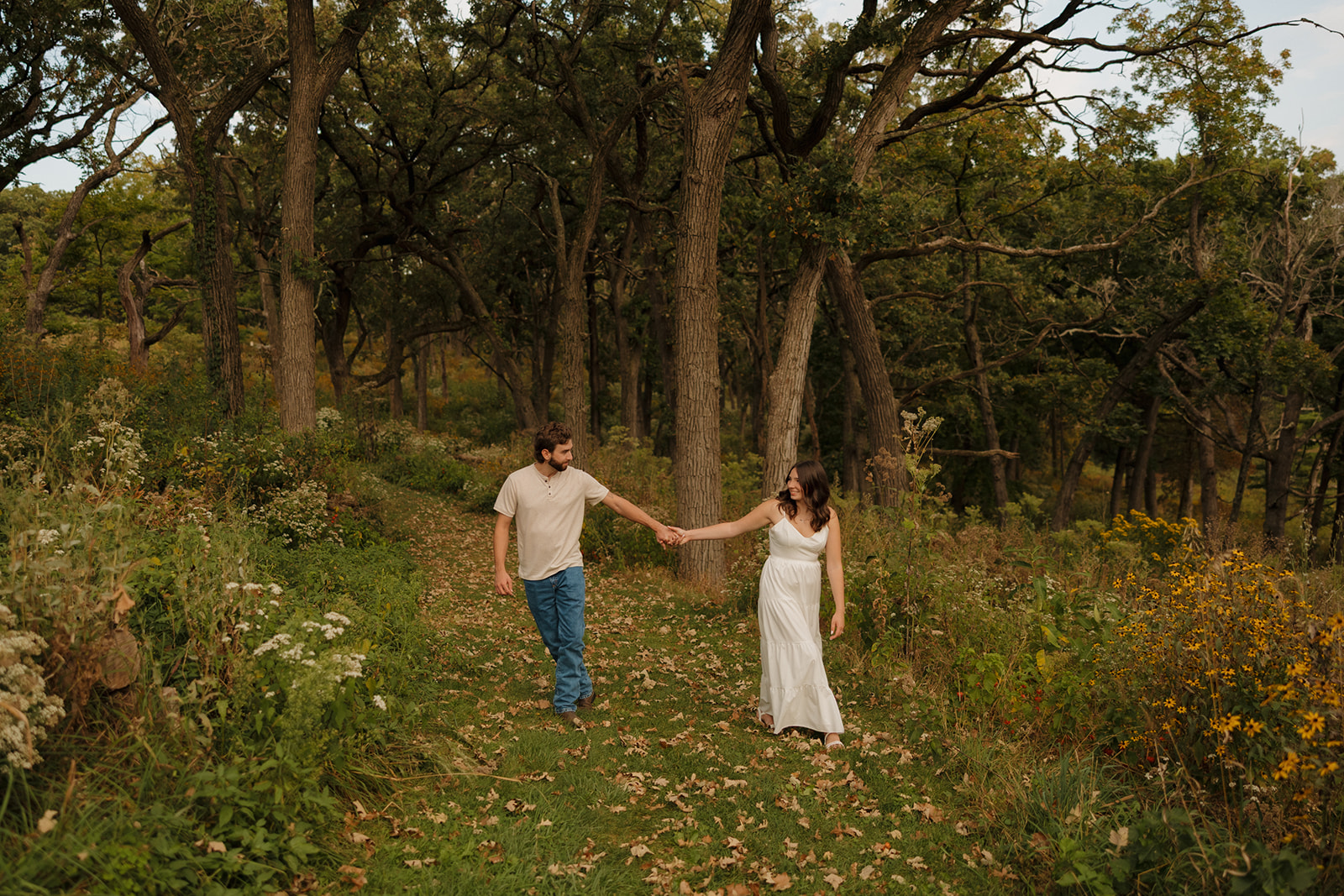 Couple walking hand-in-hand through a wooded path near Madison WI, surrounded by tall trees and wildflowers.
