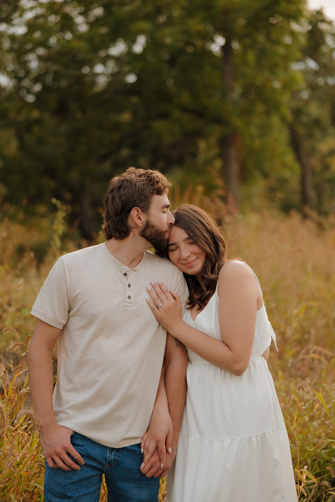 Man kisses his fiancée’s forehead while she leans into him with eyes closed, standing in a quiet field near Madison WI.