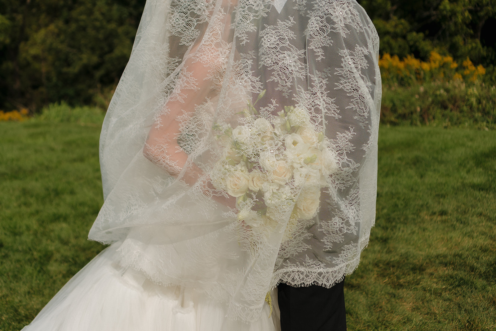 Close-up of bride and groom wrapped in her lace veil, soft white bouquet tucked between them—a dreamy, intimate moment.