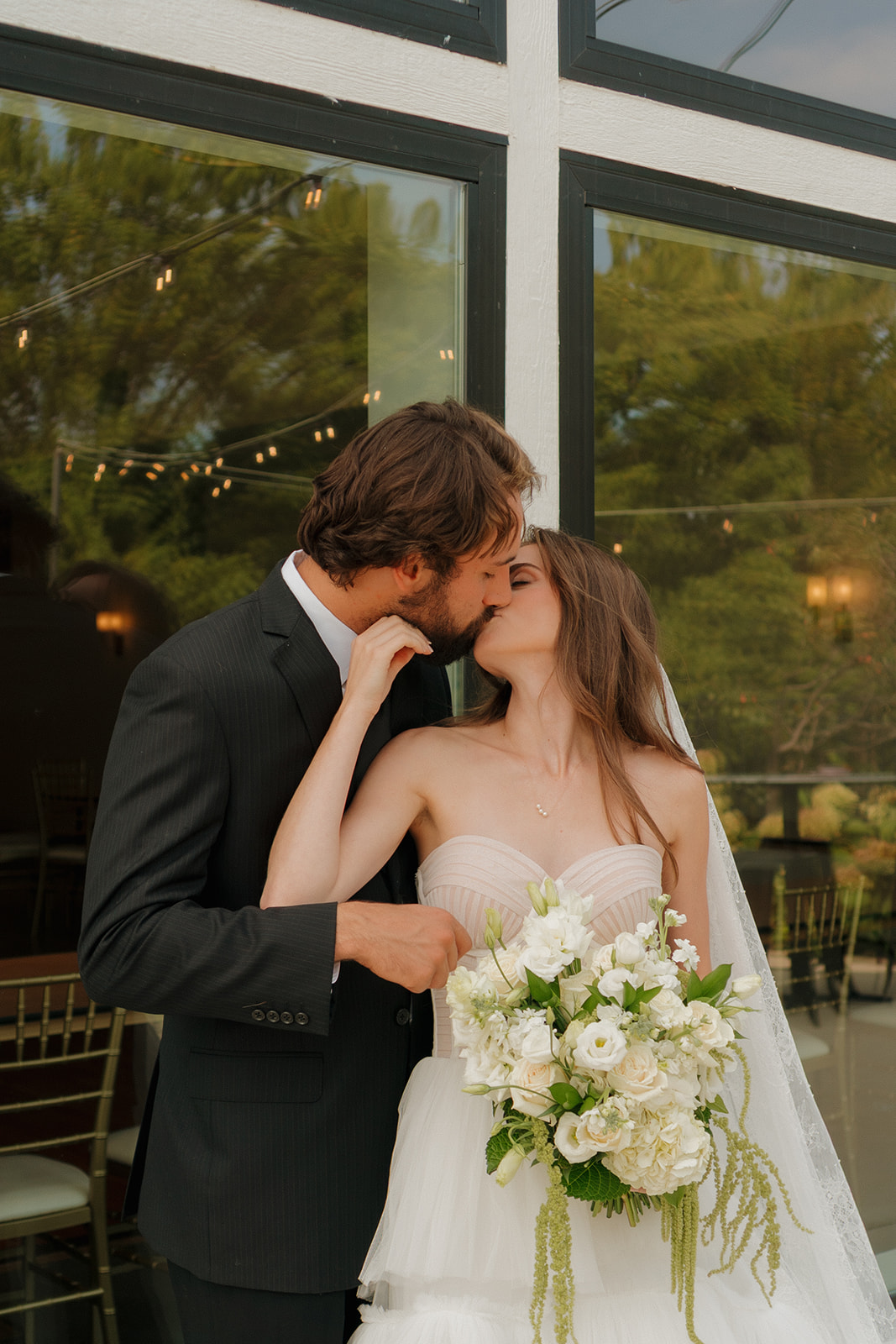 Newlyweds share a sweet kiss under soft string lights, bride holding a romantic white bouquet—picture-perfect for weddings in Wisconsin.
