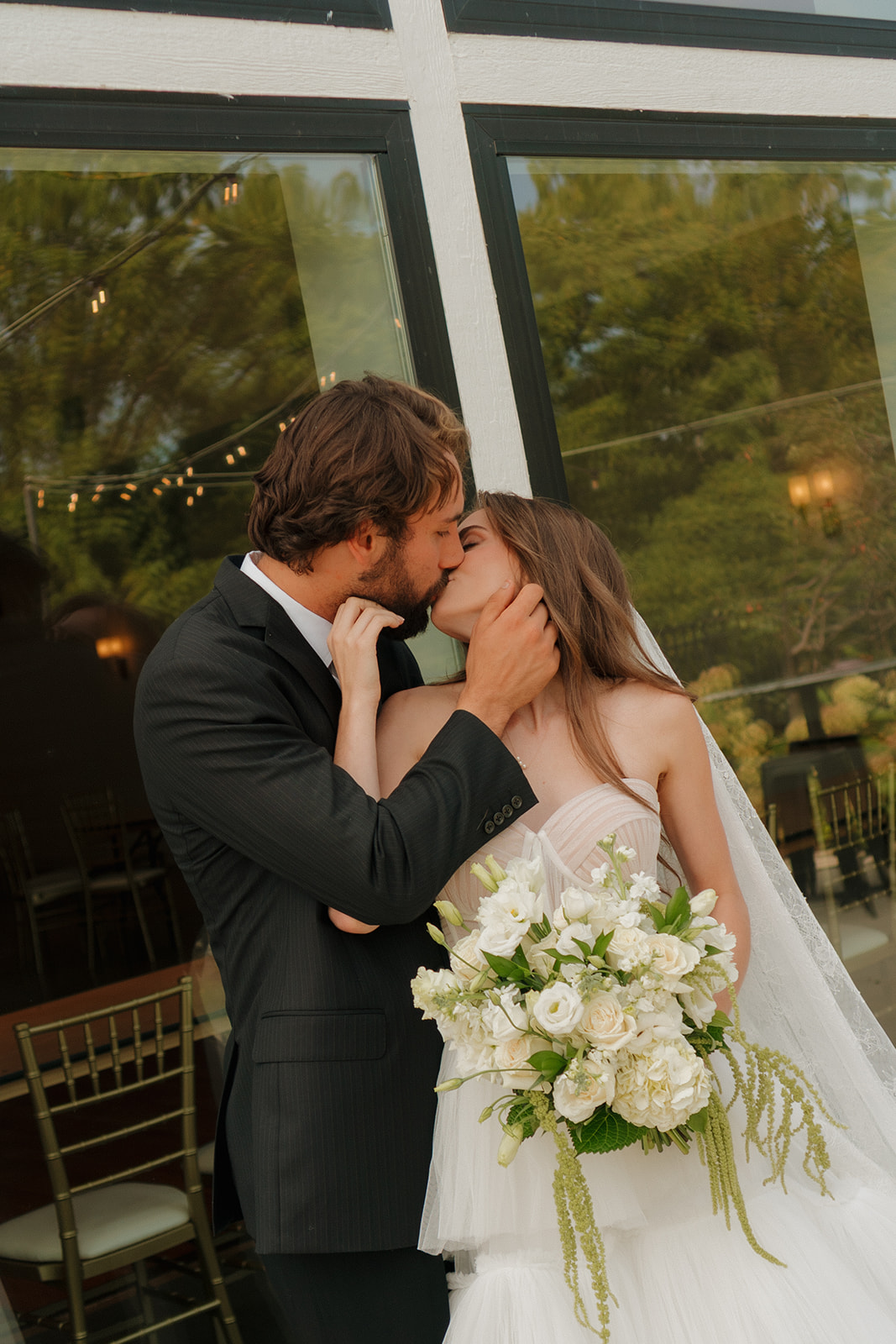 Newlyweds sharing a romantic kiss outside their venue, bride holding a cascading bouquet of white blooms and greenery.