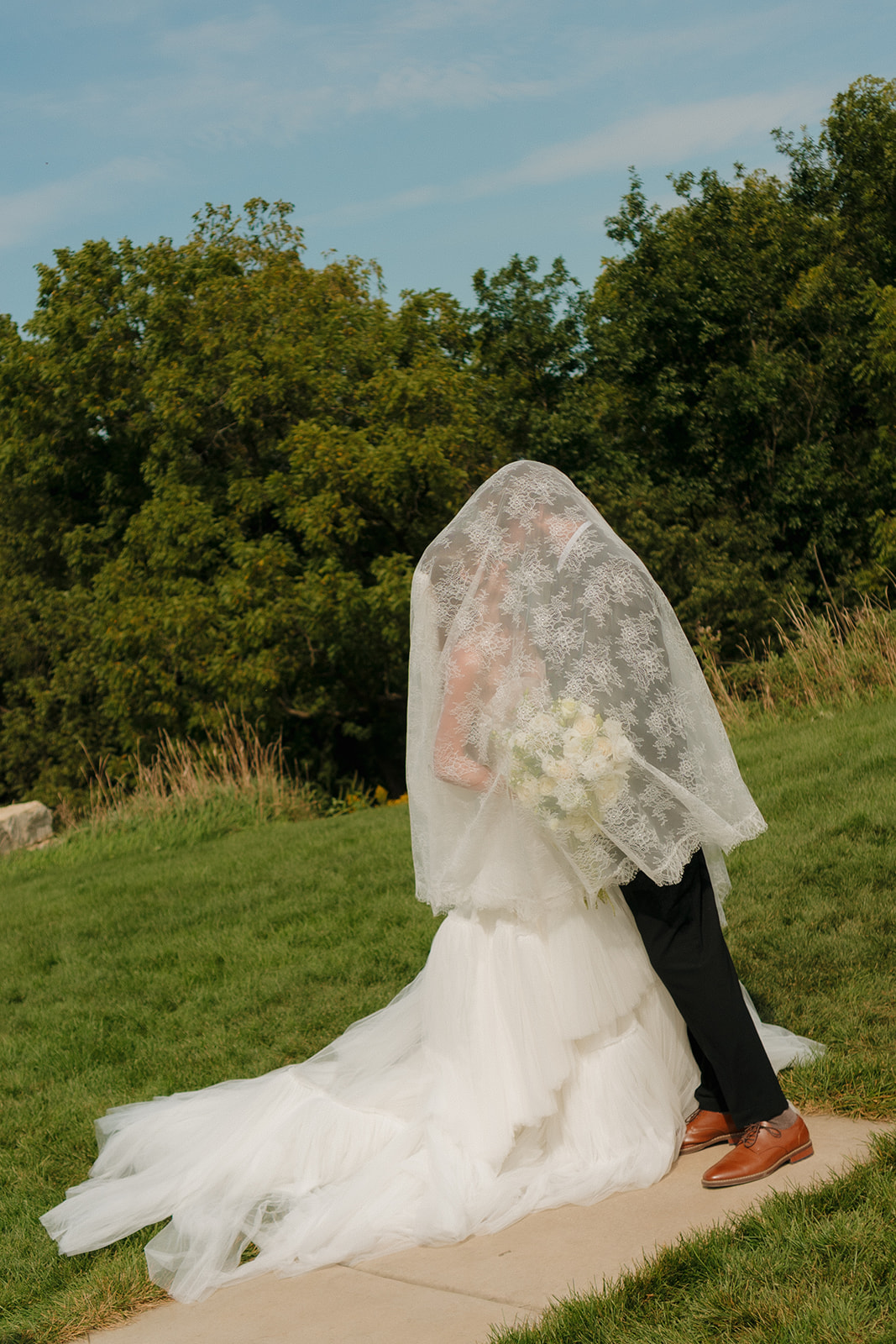 Playful, intimate moment as the couple hides under the bride’s lace veil during golden hour, standing on a path with lush greenery behind them.