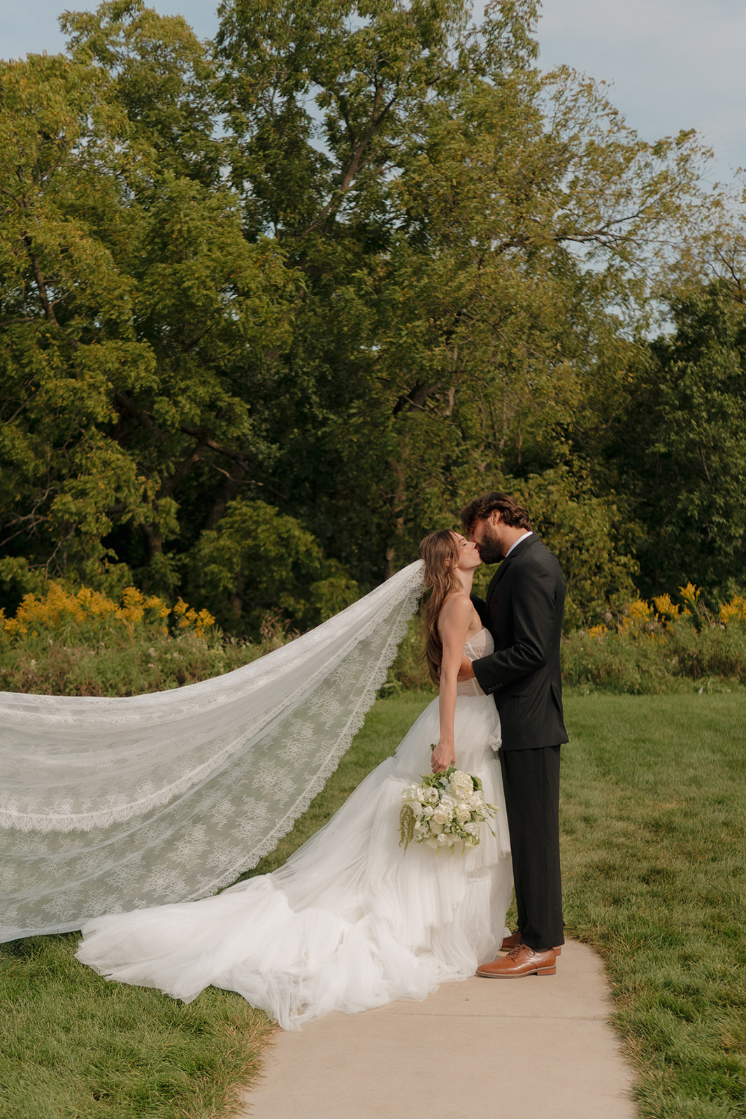 Bride and groom sharing a kiss on a quiet path, her lace veil flowing behind them—a romantic moment from weddings in Wisconsin.