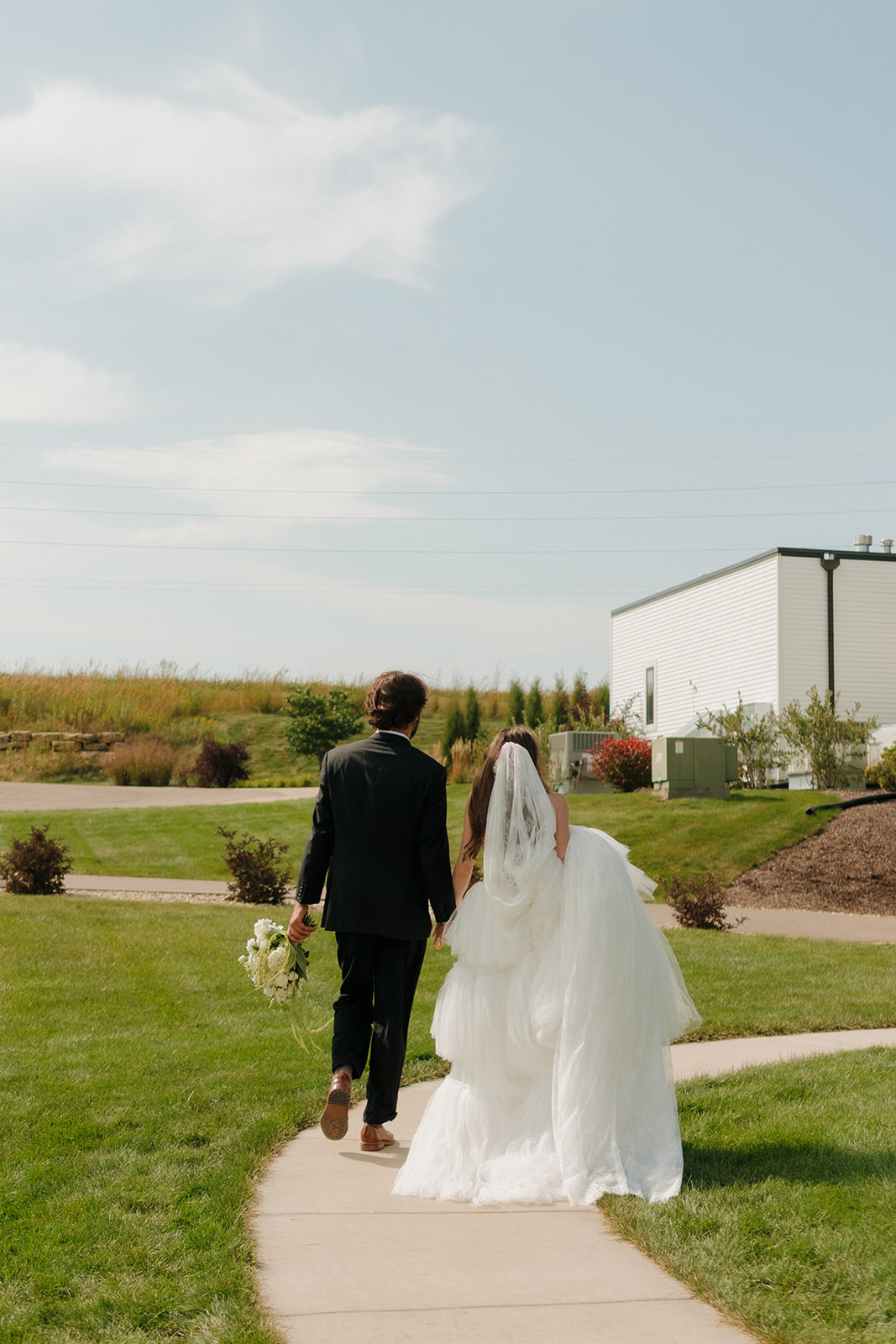 Bride and groom walk hand-in-hand down a winding path under a clear sky, bouquet trailing behind—a peaceful scene from weddings in Wisconsin.