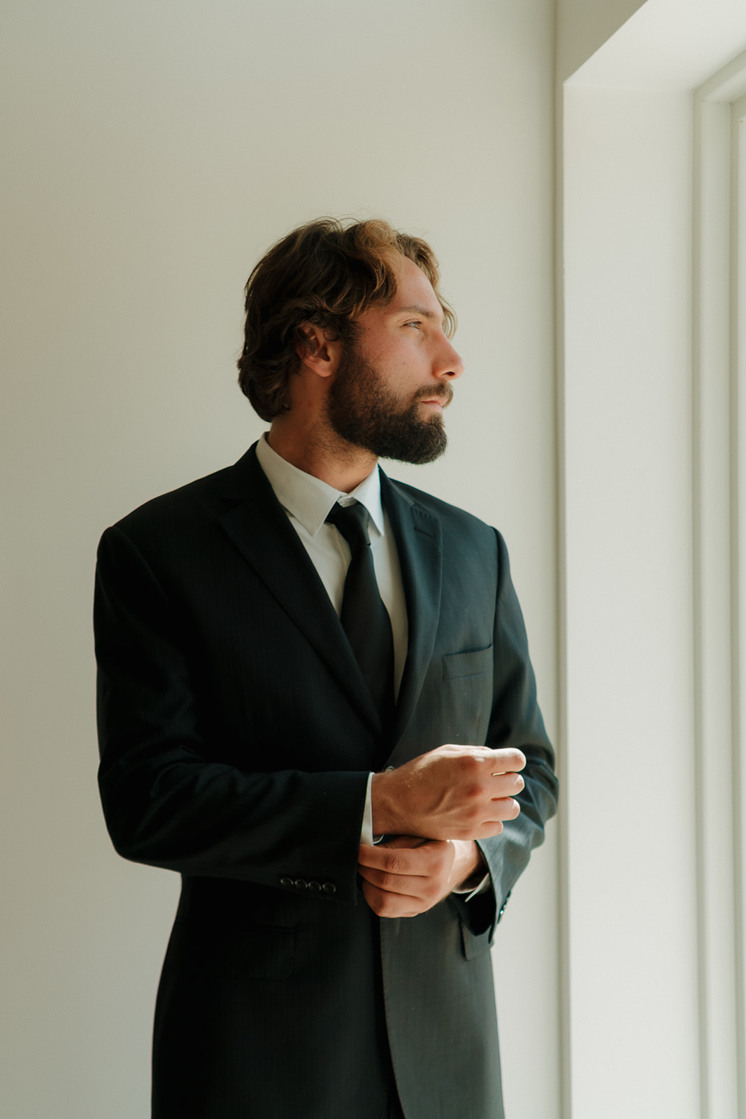Groom adjusts his cuff with a quiet confidence, standing in moody natural light by the window in a classic black suit.