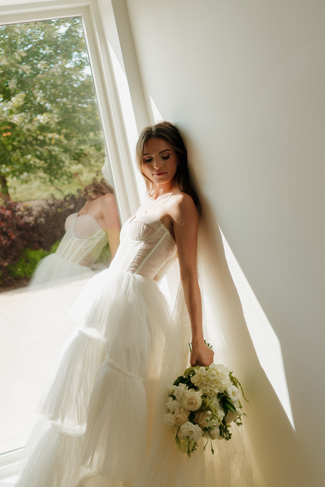 Bride stands effortlessly radiant in a sun-drenched corner, bouquet in hand and layers of her gown catching the light.