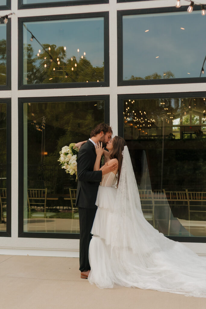 Bride and groom share a kiss outside their venue, floor-to-ceiling windows and twinkle lights reflecting the beauty of weddings in Wisconsin.