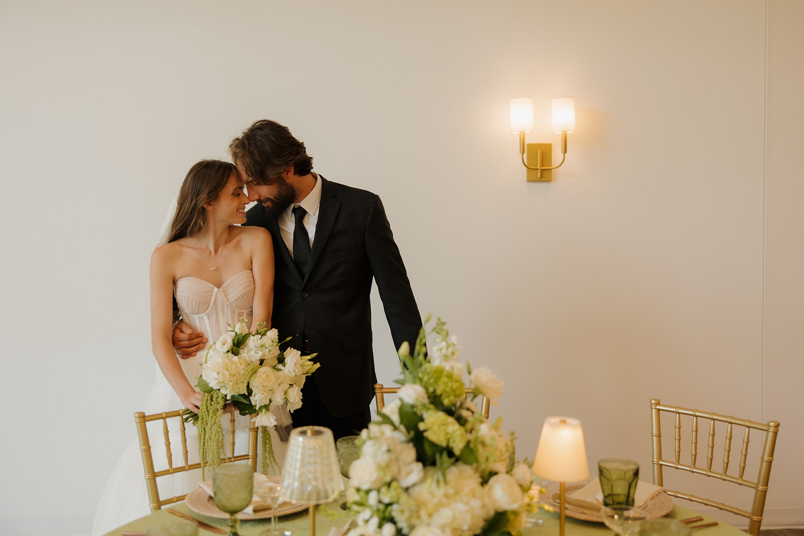 Bride and groom sharing a forehead-to-forehead smile behind their candlelit sweetheart table, surrounded by white florals and gold chairs.