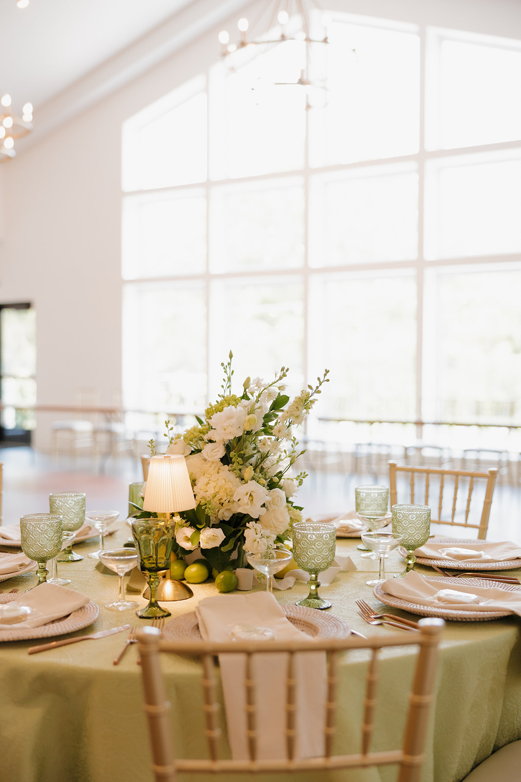 Refined reception setup with green glassware, soft white blooms, and a glowing lamp centerpiece—all set against floor-to-ceiling windows.