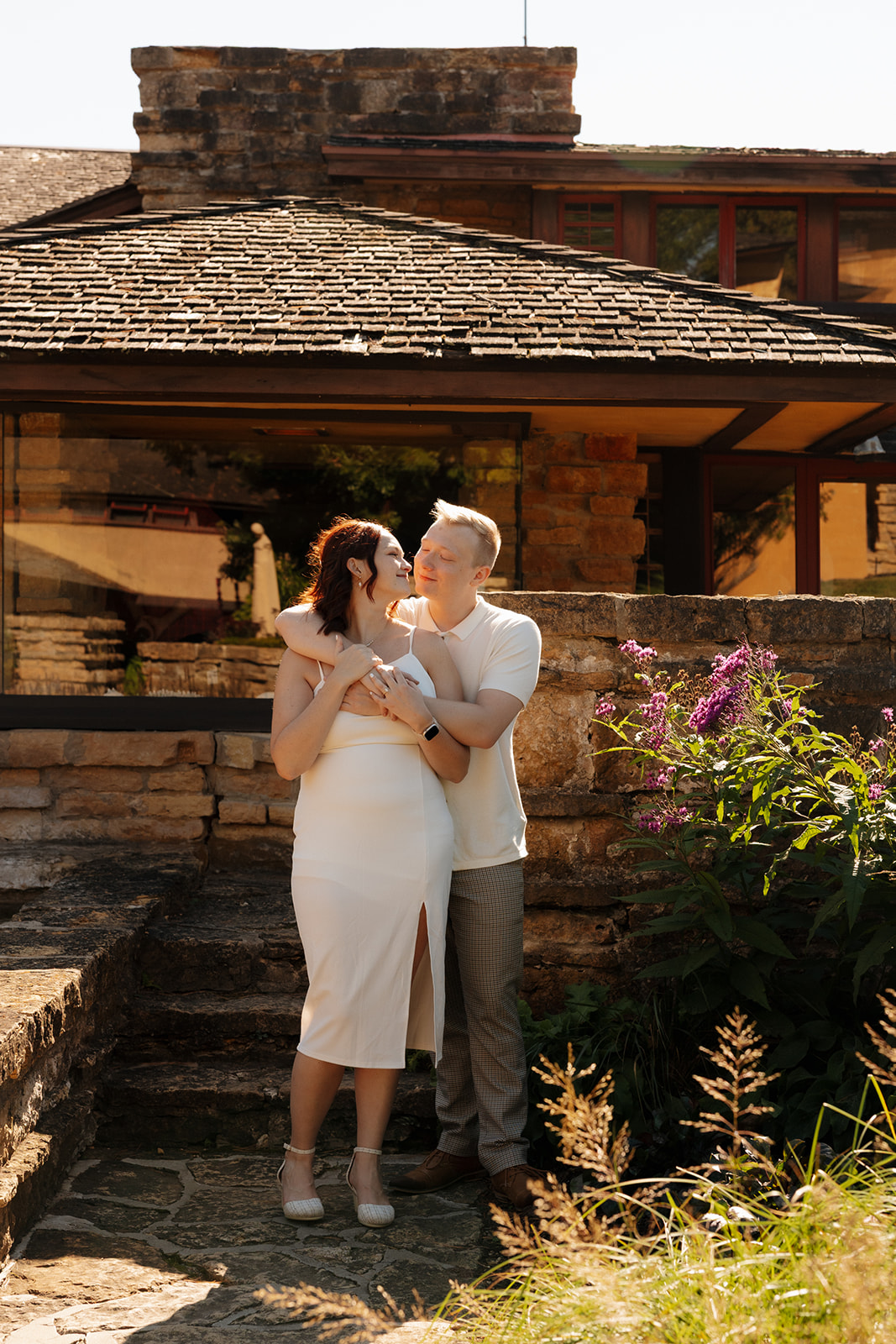 Wrapped in an embrace on stone steps, the couple shares a playful look beneath blooming purple flowers and a low-slung roofline.