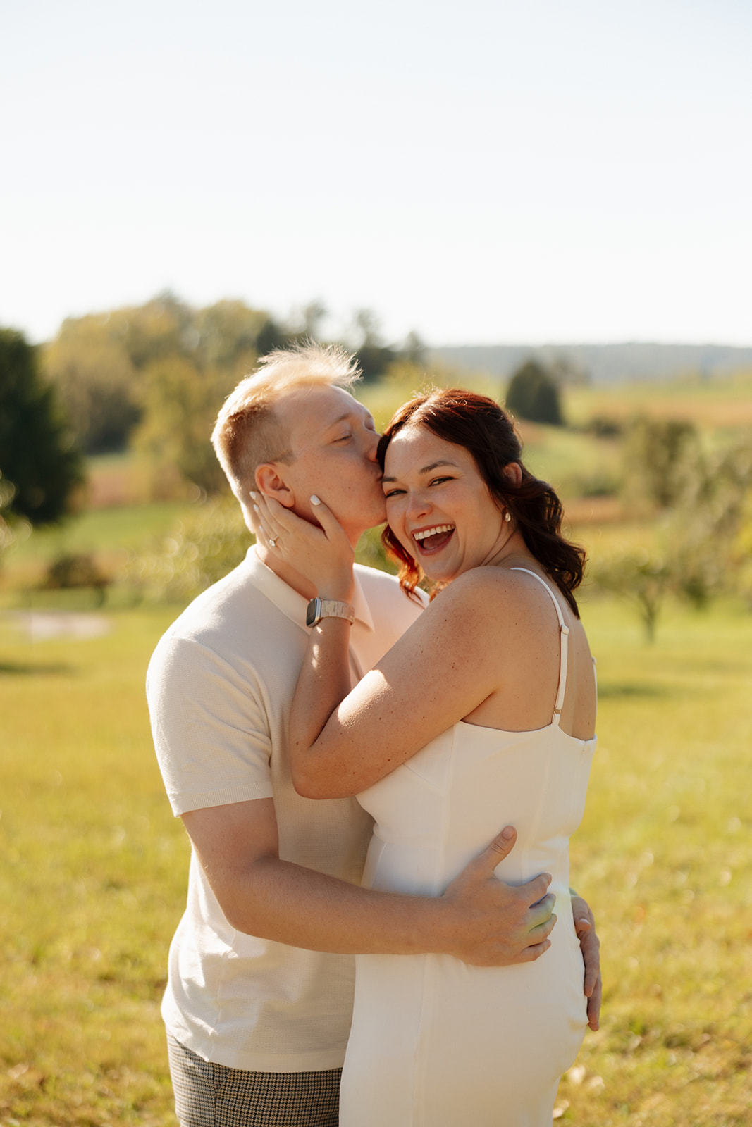 Groom-to-be kisses bride’s cheek as she beams with joy in a sunlit field, capturing laughter, love, and carefree connection.