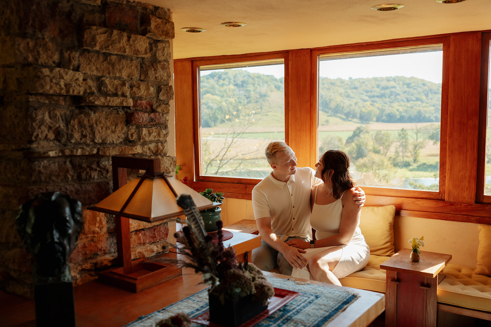 Couple cuddled up on a built-in bench in a sunlit room, framed by large windows overlooking rolling green hills—an intimate and scenic idea for engagement photos with an architectural twist.