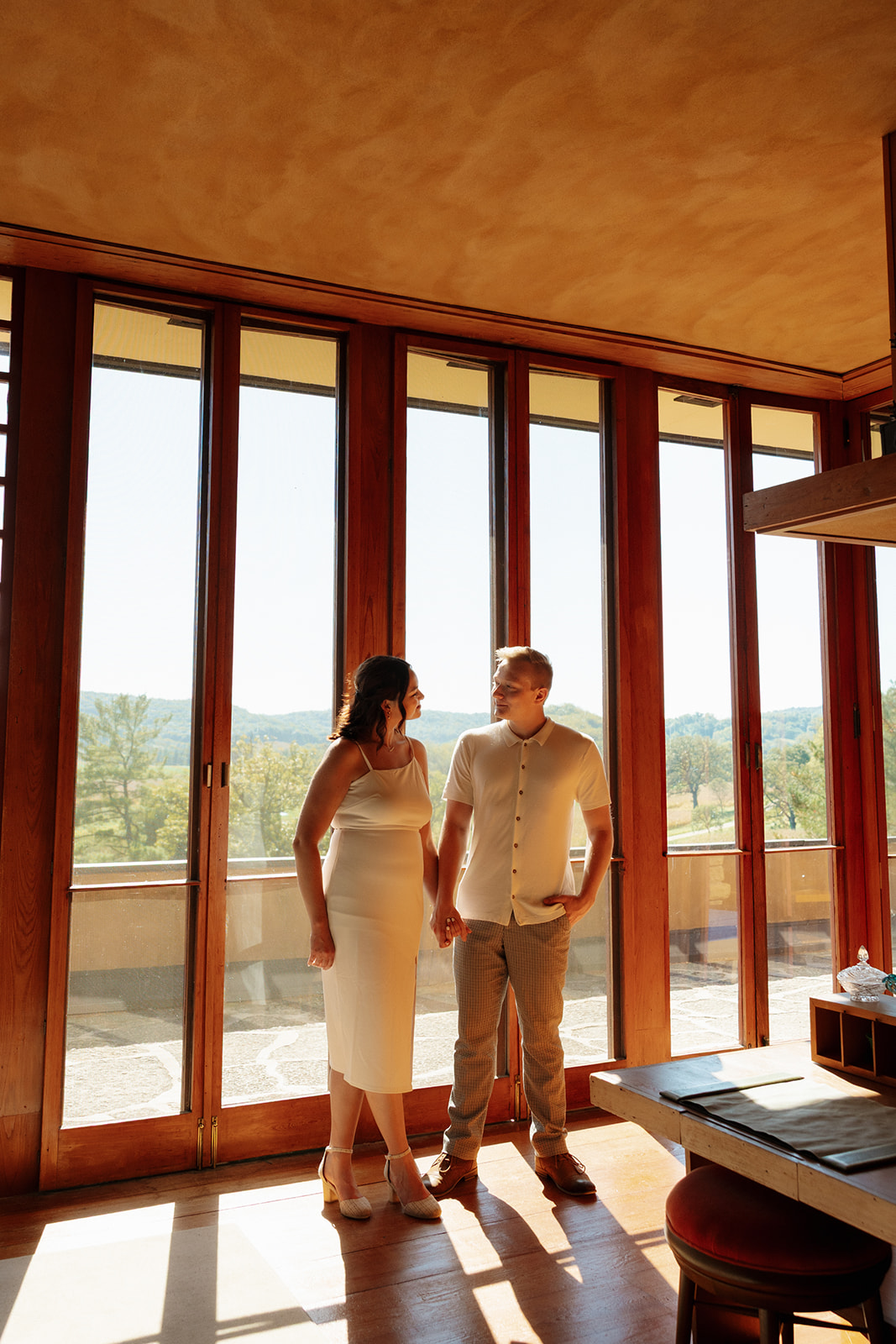 Holding hands inside a modern sunlit space with towering windows, the couple locks eyes in a calm and connected moment.