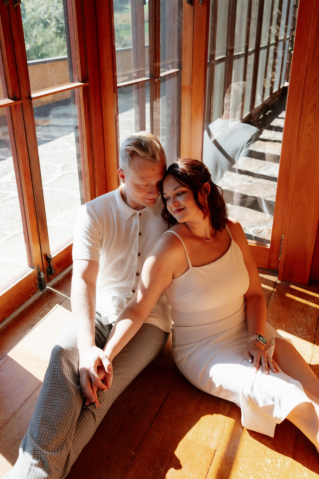 Couple sitting together on the wooden floor in golden sunlight, leaning into each other with soft smiles in a cozy, glass-walled space.
