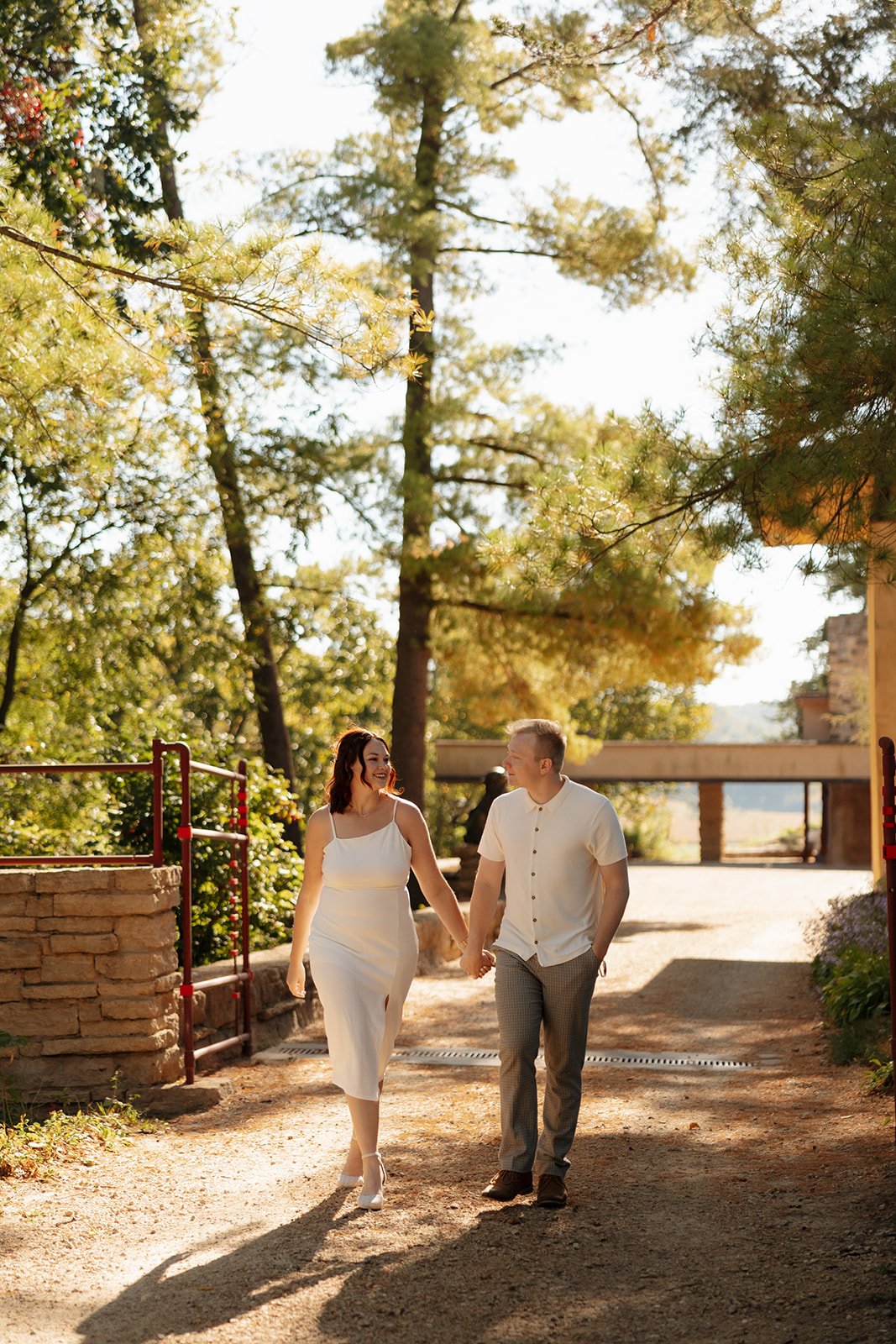 Hand in hand, walking a sun-kissed path between tall trees and stonework, the couple looks completely in sync—a simple yet heartfelt idea for engagement photos.
