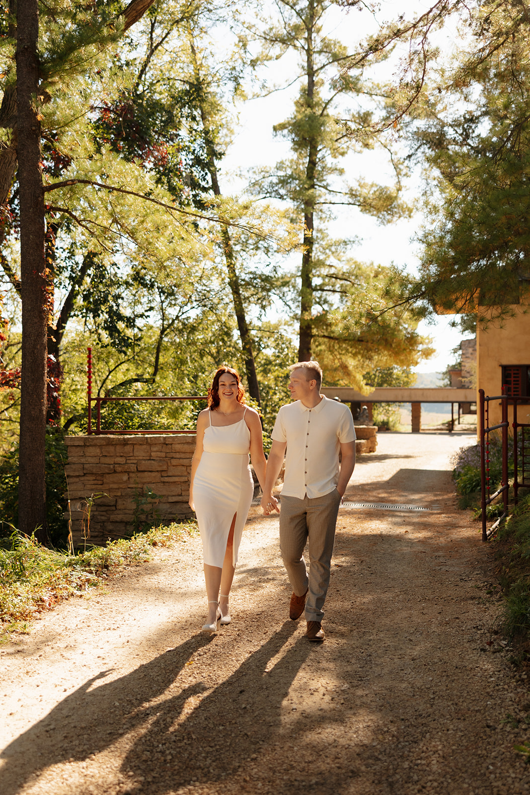 Walking hand in hand down a sun-dappled path, the couple smiles at each other, surrounded by tall trees and soft shadows.