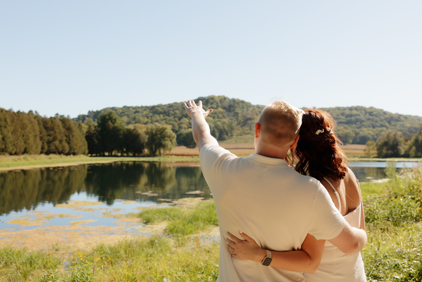 From behind, the couple embraces by a peaceful lake as one partner gestures toward the hills, soaking in the view and dreaming out loud.