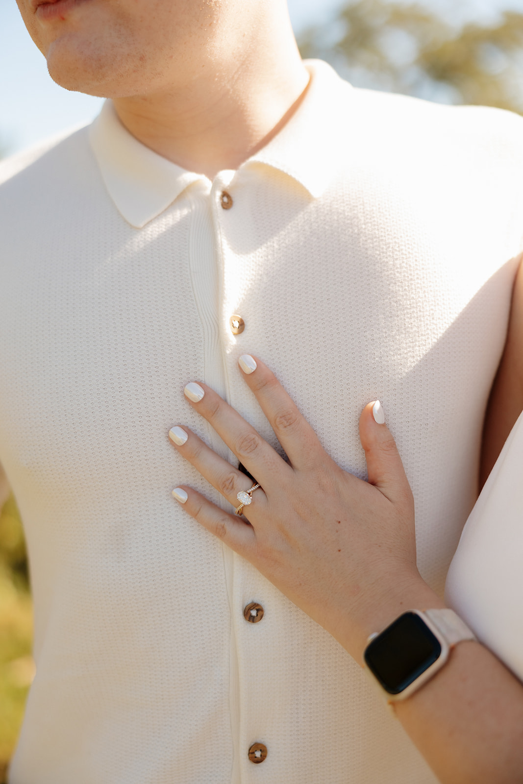 Close-up of a hand resting on the groom’s chest, showcasing a radiant engagement ring and fresh white manicure against a soft cream shirt.