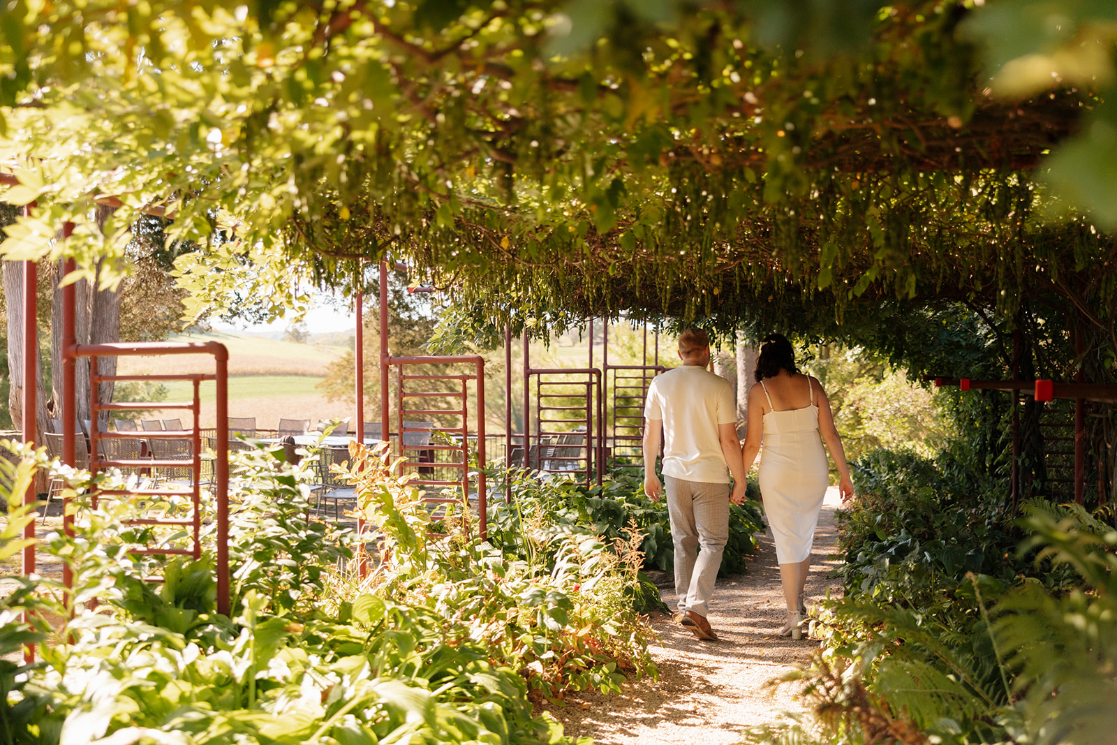 Walking away down a lush garden path hand in hand, the couple disappears into a dreamy green tunnel—an intimate and whimsical idea for engagement photos with movement and depth.