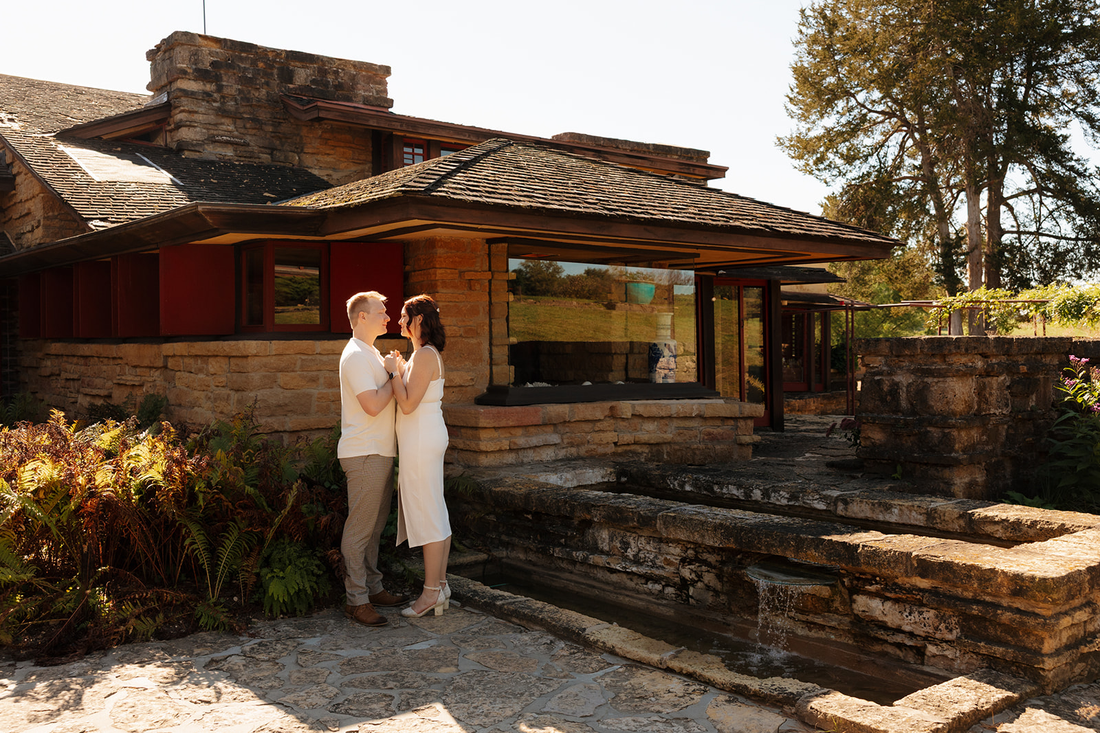 Couple standing close outside a Frank Lloyd Wright-style home, surrounded by warm stone, sunlight, and architectural charm—an elegant idea for engagement photos with timeless character.