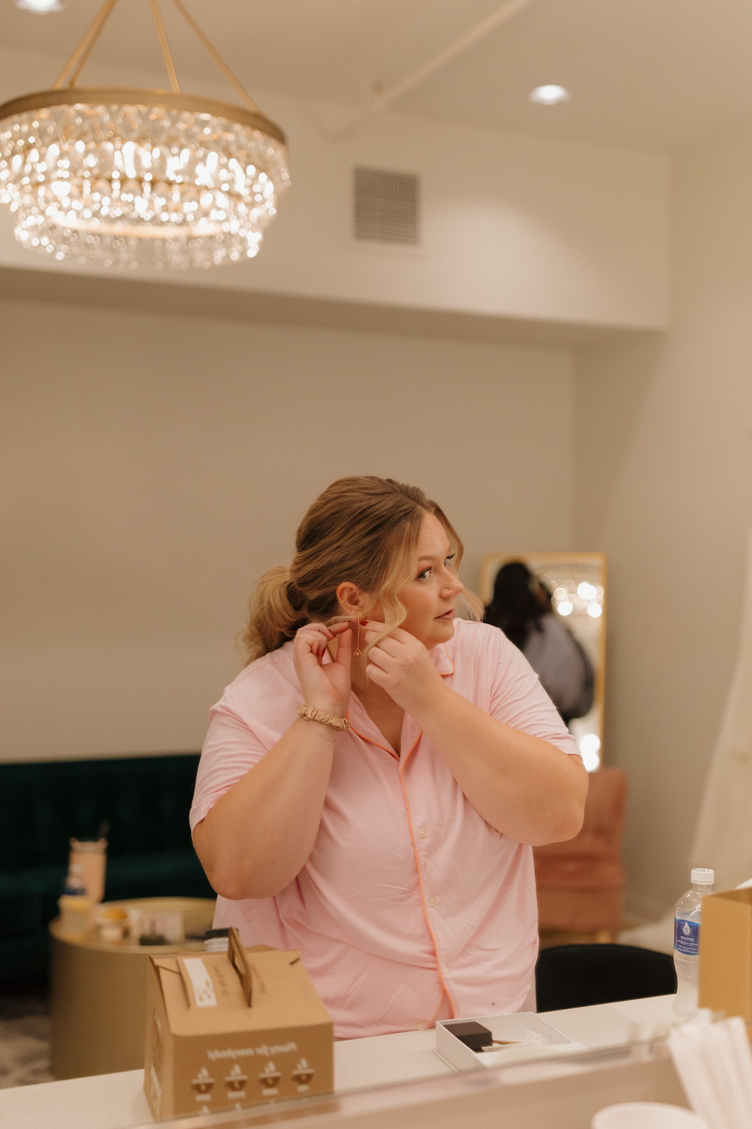 Bride getting ready and putting on earrings in a glam bridal suite with a crystal chandelier and soft lighting.