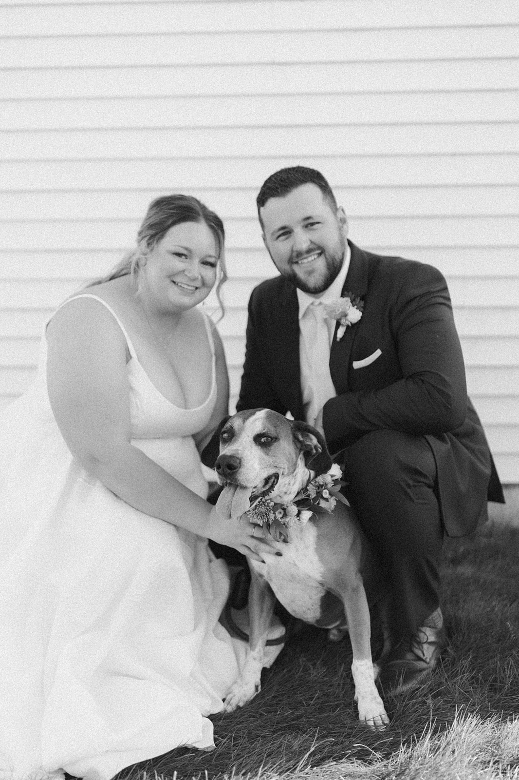 Black and white portrait of a couple posing with their dog outside their Wisconsin wedding venue.