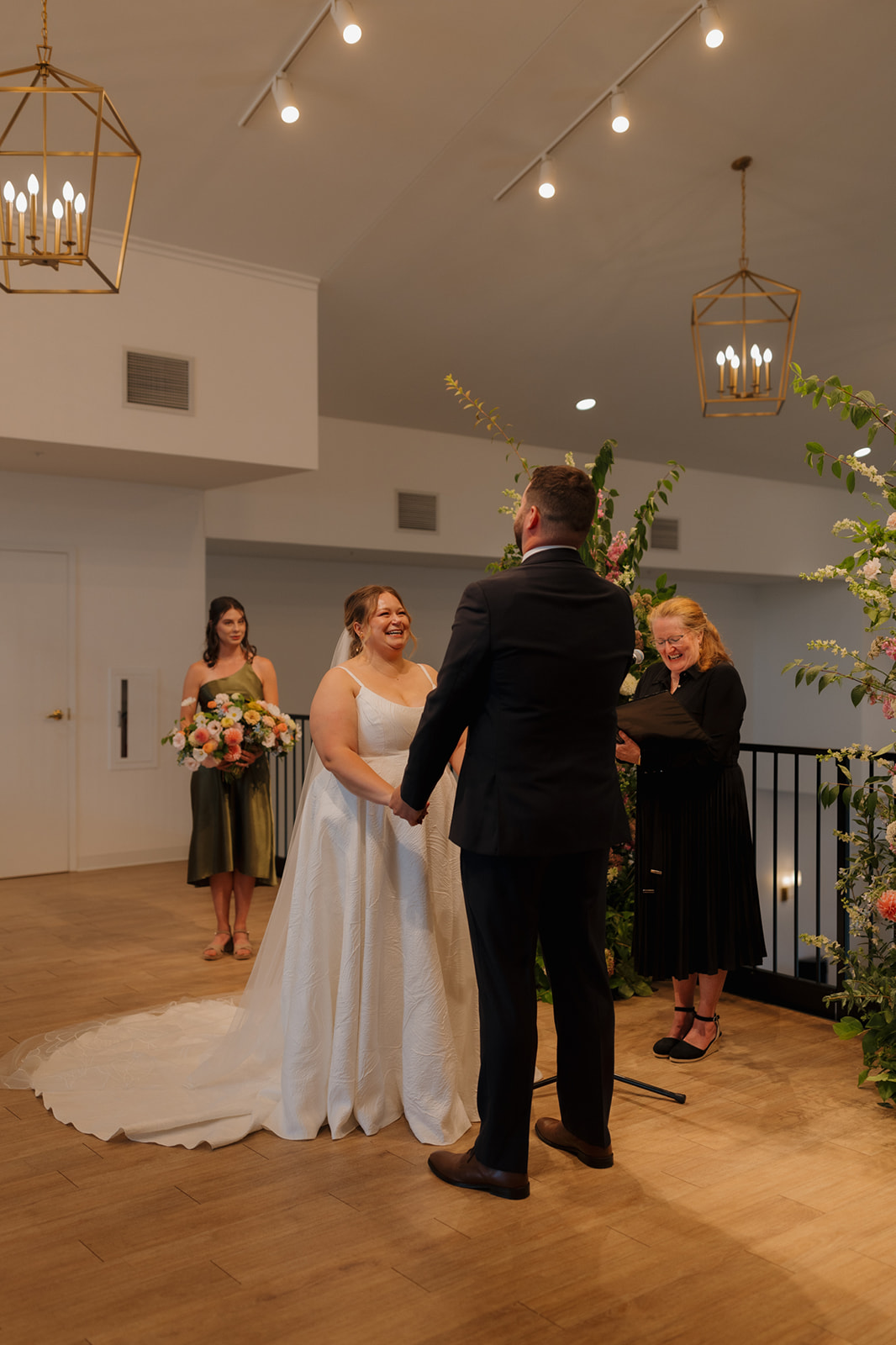 Bride and groom holding hands during their ceremony, framed by floral pillars and soft light in a modern indoor space.