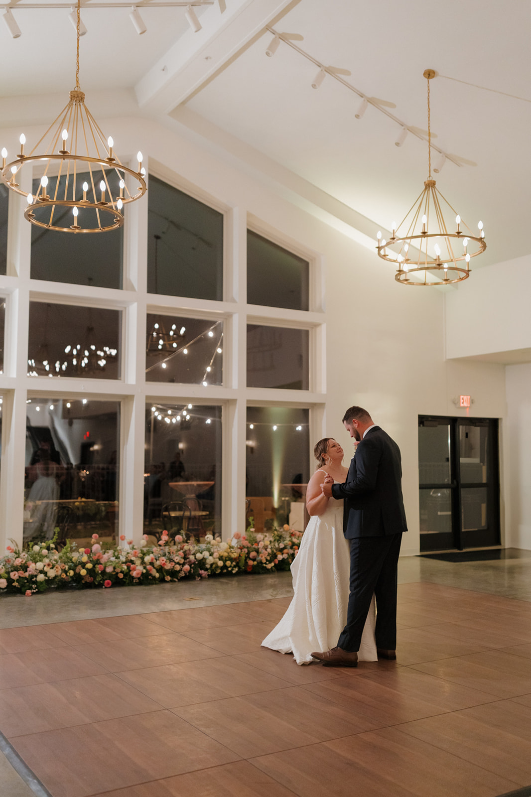 Romantic first dance beneath chandeliers, framed by tall windows and floral arrangements at a wedding venue in Wisconsin.