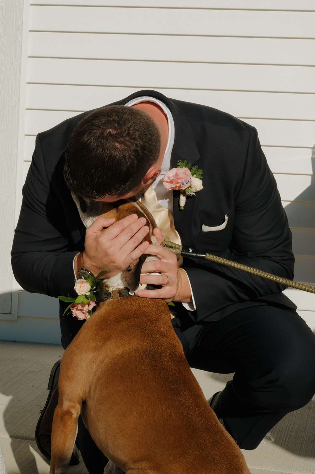 Groom cuddles his dog while kneeling in front of white siding in golden afternoon light.