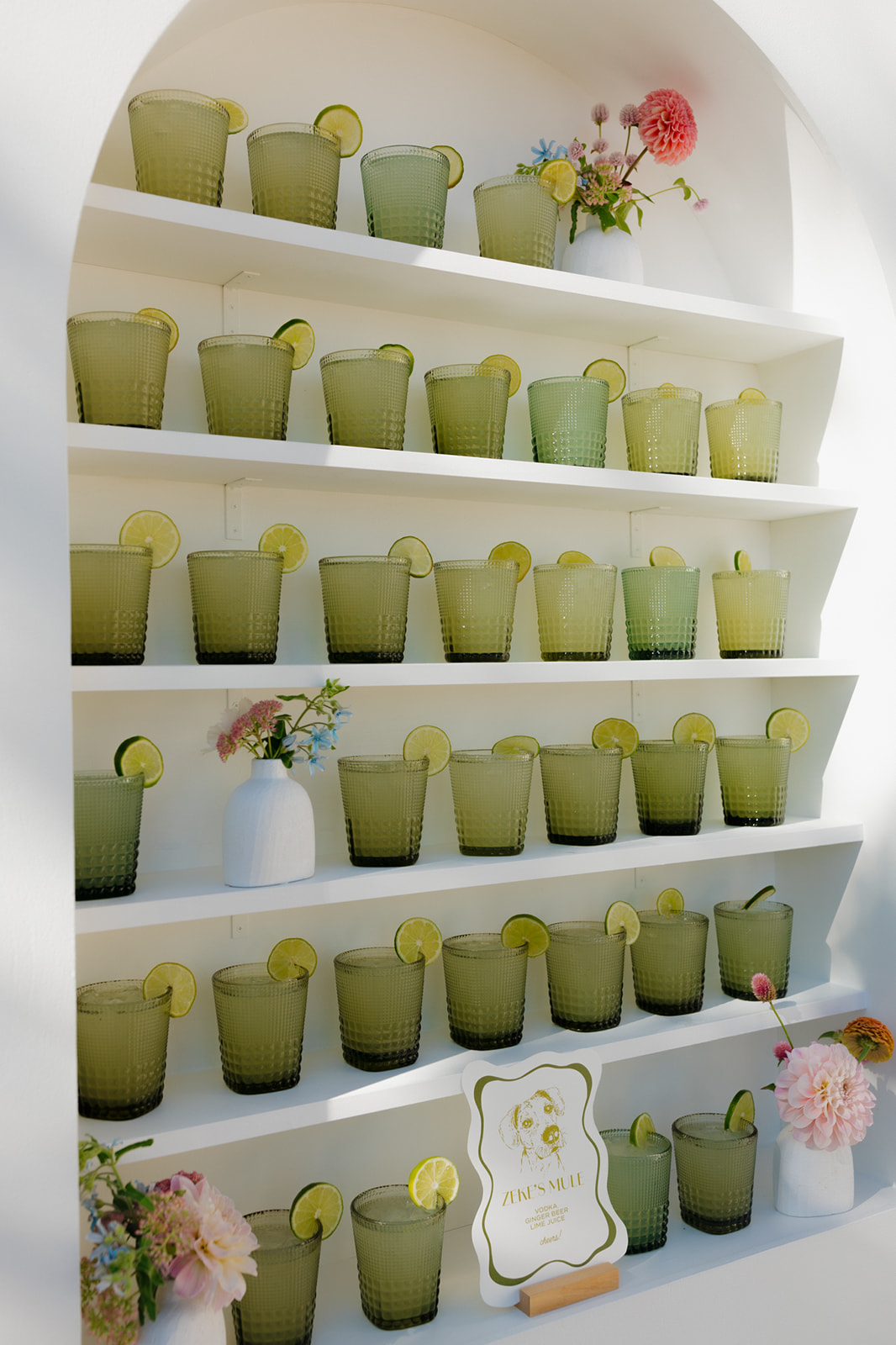 Rows of green signature cocktails with lime garnishes displayed on white shelves, styled with florals for a modern wedding in Wisconsin.