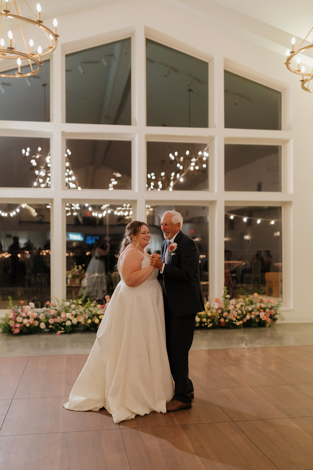 Bride shares a joyful dance with her father in front of floor-to-ceiling windows and florals.