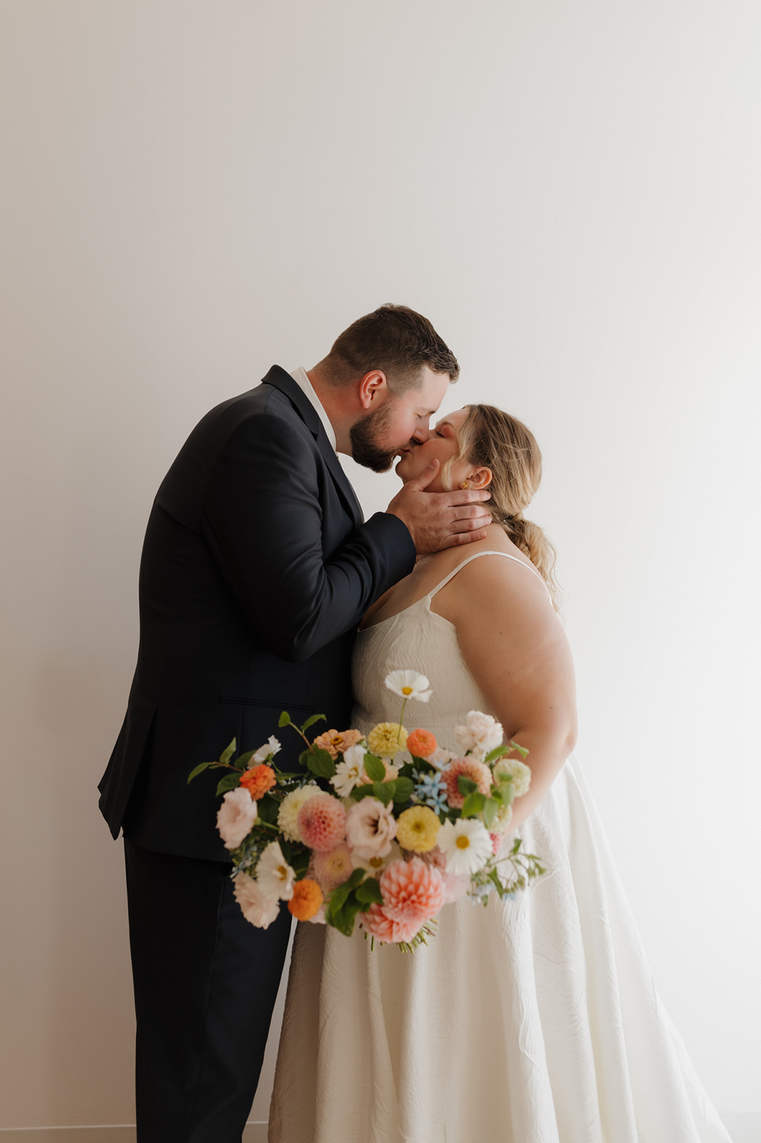 Groom kisses the bride while she holds a lush, pastel bouquet inside a modern white space.
