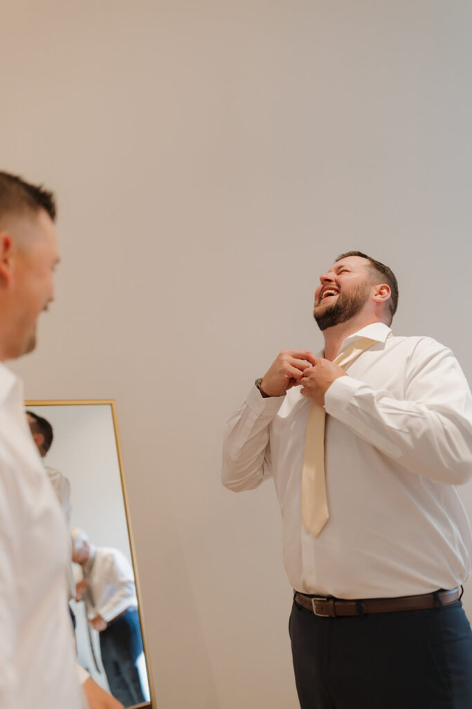 Groom laughing mid-tie, sharing a joyful pre-ceremony moment with his best man.