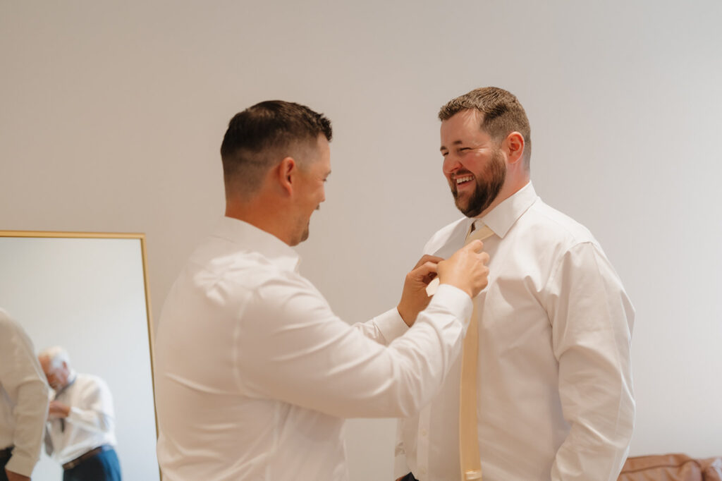 Groom getting ready with a friend’s help, both smiling and relaxed in a modern suite at a wedding venue in Wisconsin.