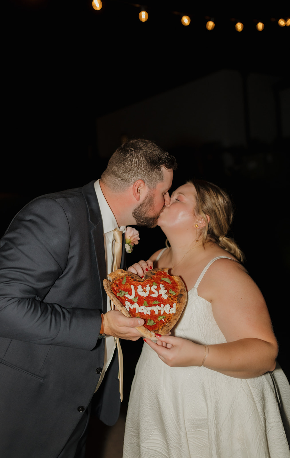 Newlyweds share a kiss while holding a heart-shaped pizza that says “Just Married.”