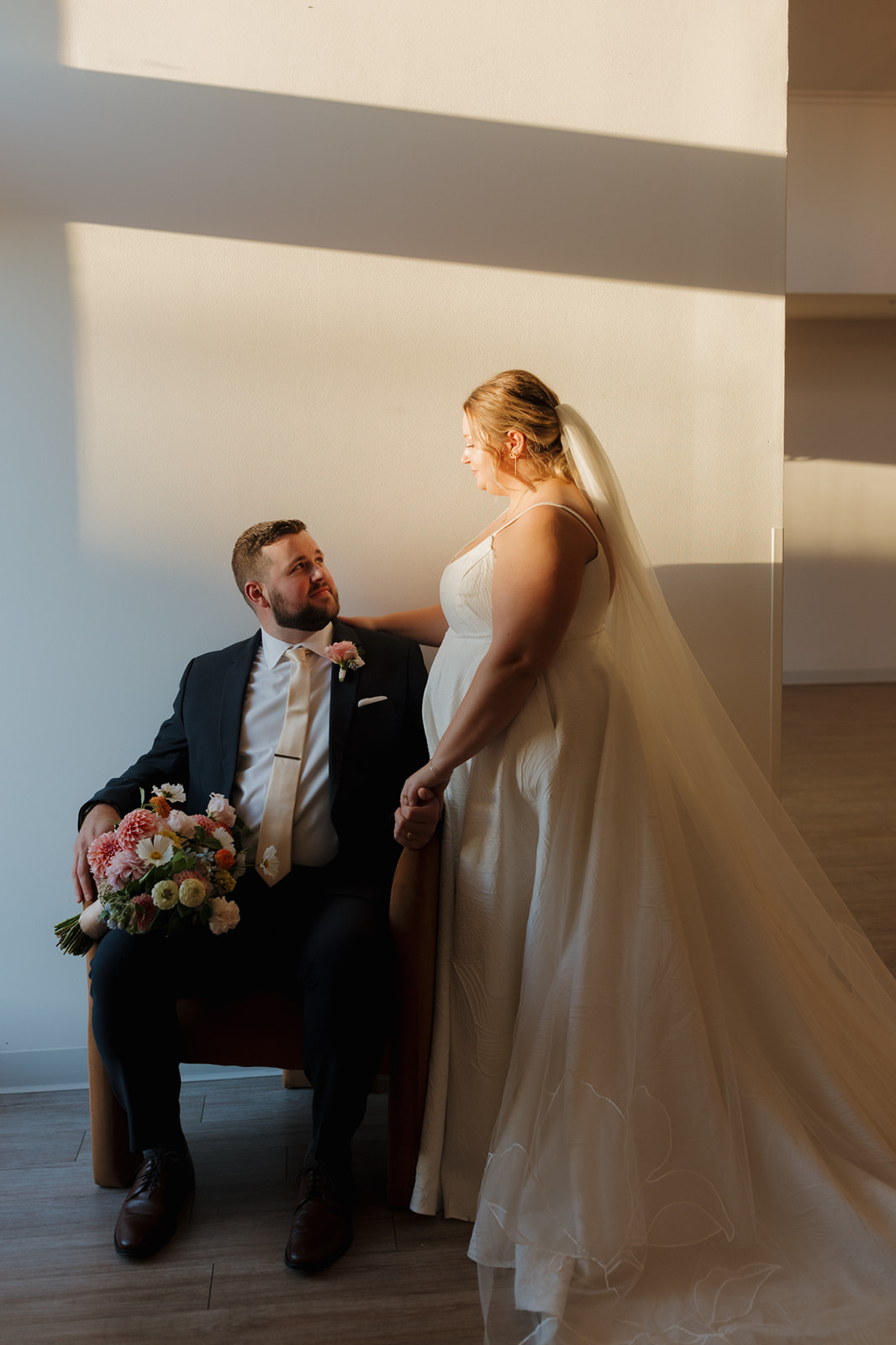 Bride and groom sharing a tender moment as golden hour light spills across the room, highlighting the textures of her dress and bouquet.