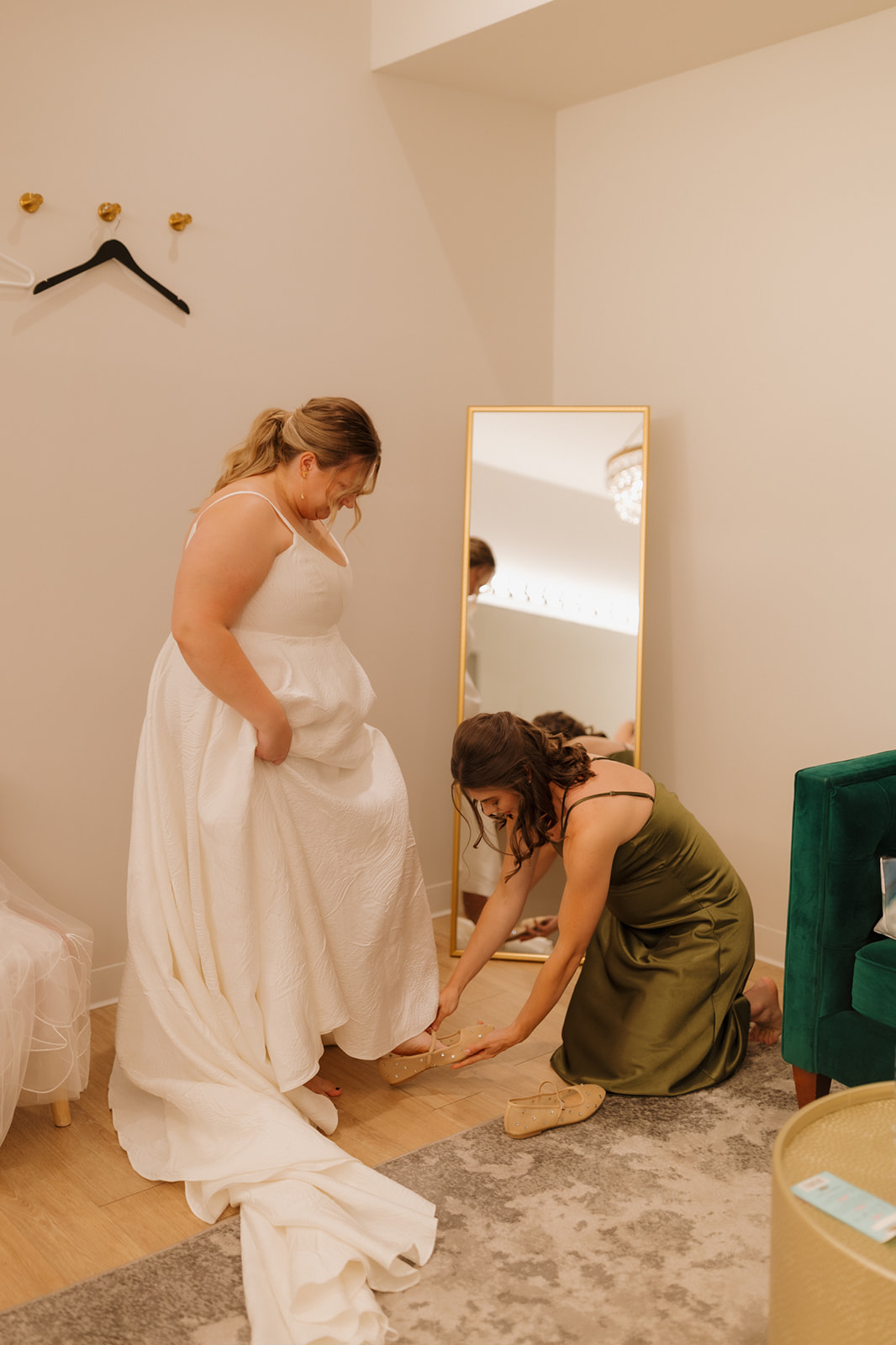 Bridesmaid helping the bride with her shoes in a cozy getting-ready suite, with a mirror and velvet chair nearby.