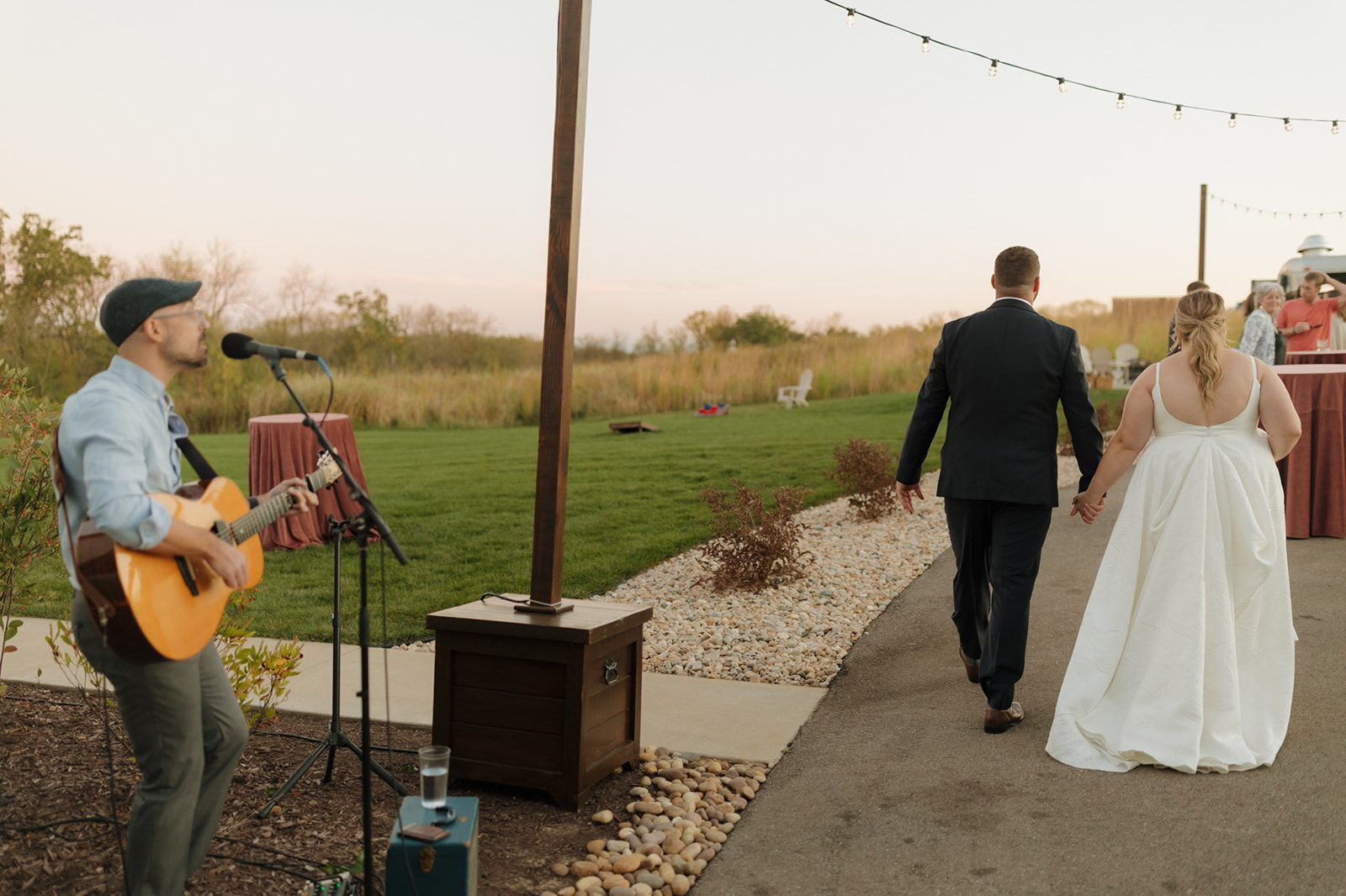 Bride and groom walking hand-in-hand toward their reception while a live musician plays acoustic guitar at golden hour.